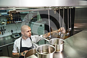 Cooking soup. Young male chef in apron choosing a ladle for his famous soup while standing in a restaurant kitchen