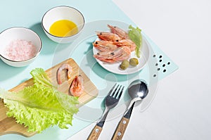 Cooking of spring salad with shrimps, green olive, greens, olive oil, ingredients on pastel minty background with shadow in hard.