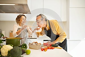 Cooking together concept. Senior woman feeding her husband while preparing dinner in kitchen, making delicious meal