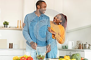 Cooking together. Happy loving black wife hugging husband while man cooking salad for dinner in kitchen at home