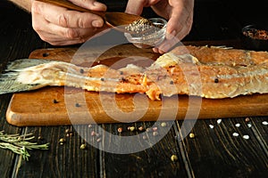 Cooking trout fish on the kitchen table. A cook adds coriander to a fish steak with a spoon before salting or canning