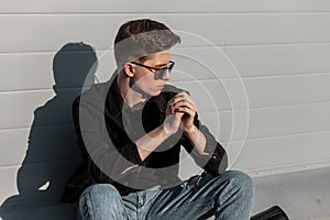 Cool American young hipster man in fashionable sunglasses in stylish casual denim clothes with hairstyle is rests near building on