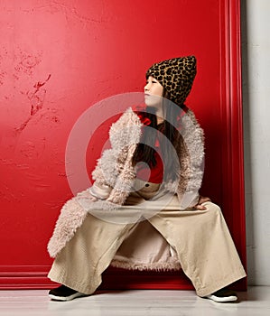 Cool asian kid girl in furcoat and wide pants is posing with her legs wide apart and hands on her knees looking aside