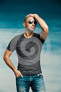 Cool bald man in a pilot`s sunglasses posing in a gray T-shirt holding his hand in his pocket