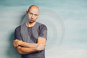 A cool bald man standing with his back against blue wall arms crossed on his chest, looking to the side and smiling