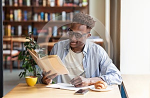 Cool black guy reading book and taking notes during his studies at cafe