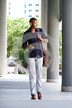 Cool black guy walking in the city with mobile phone and bag