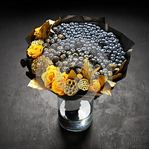 Cool bouquet of blueberries and blackberries decorated with yellow roses and gold leaves standing in a vase on a black background