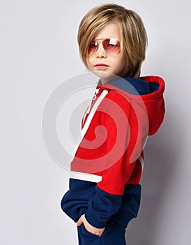 Cool boy in aviator sunglasses in blue and red hoodie with long hair posing on gray