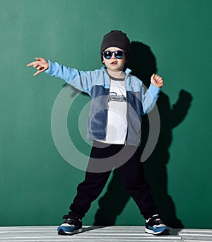 Cool boy in blue sunglasses, headwear, fleece jacket, pants and sneakers is showing rock sign on green