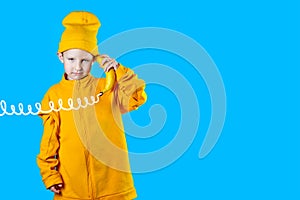 Cool boy in a bright yellow jacket and hat holds a banana in his hands like a phone. On blue background