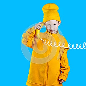 Cool boy in a bright yellow jacket and hat holds a banana in his hands like a phone. On blue background