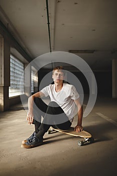 Cool boy with brown hair sitting on skateboard and dreamily looking in camera. Young thoughtful man in white t-shirt