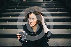 Cool brunette girl holding camera sitting on stair inside the building. Woman photographer