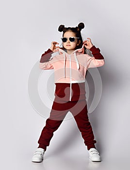 Cool brunette kid girl in modern fashion pink brown sportwear and sunglasses stands holding cowl of her hoodie