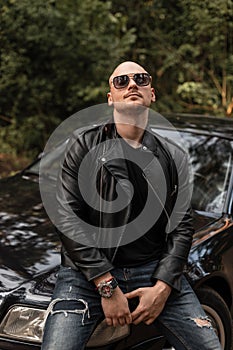 Cool brutal bald man with beard in fashion sunglasses in stylish vintage black jacket sits on black modern car outdoors at sunset