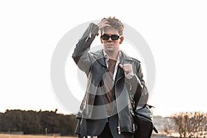 Cool brutal young man hipster in vintage sunglasses in fashion oversized black leather jacket in a shirt straightens stylish