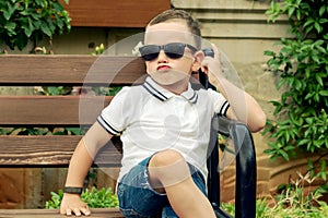 A cool child in sunglasses sits on a bench with his legs crossed.