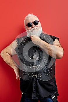 Cool elderly gray-haired mustache bearded man in black leather jacket posing isolated on red wall background