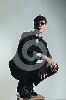 cool elegant best man with glasses wearing black tuxedo and crouching