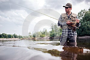 Cool fisherman stands in water and looks straight forward. He is serious. Guy holds fly rod and wooden box of artificial