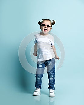 Cool frolic little baby kid girl with funny buns with bows and in sunglasses, t-shirt and blue jeans is singing, dancing
