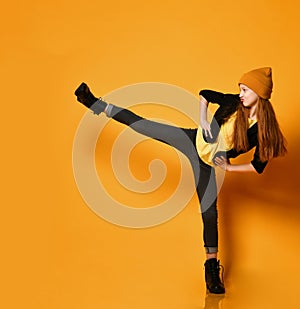 Cool and frolic red-haired girl in hat, black tight jeans and brutal shoes is kicking holding leg up high