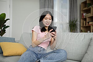 Cool gadget and application. Happy asian lady holding mobile phone, typing sms message, sitting on couch in living room