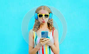 cool girl blowing red lips holding phone listening to music in wireless headphones on colorful blue