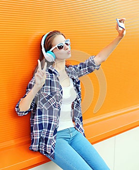 Cool girl makes self portrait on smartphone listens music in headphones over orange