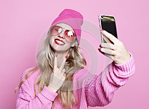 Cool girl taking photo makes self portrait on smartphone wearing pinkl clothes over pink background