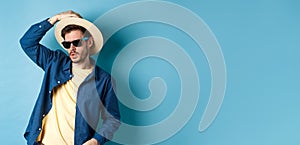 Cool guy having fun on vacation, wearing straw hat and sunglasses, looking aside sassy, standing on blue background