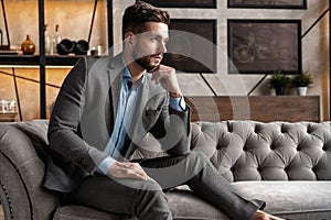 Cool and handsome. Good looking young man in full suit sitting on the sofa while spending time indoors.