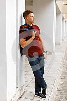 Cool handsome young man in trendy red t-shirt in a plaid fashionable shirt in blue jeans in sneakers posing on the street near a