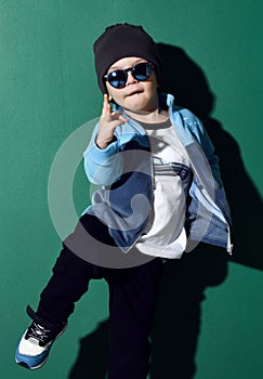 Cool kid boy in blue sunglasses, headwear, fleece jacket, pants and sneakers is dancing, showing cool sing on green