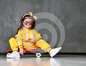 Cool kid girl in yellow hooded jumpsuit costume, sunglasses and yellow bow sits on skateboard in cool pose like just after trick