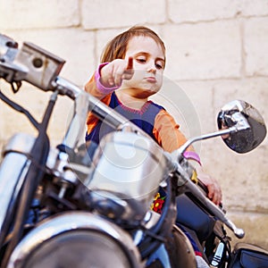Cool little biker girl playing and having fun on fashioned motorcycle. Humorous portrait of child points to the road with finger