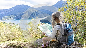 A cool little independent girl with a backpack sits in a forest on a large boulder and studies a tourist map in search of a hiking