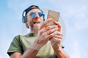 Cool man in baseball cap, wireless headphones and blue sunglasses browsing in his mp3 player playlists smartphone device and