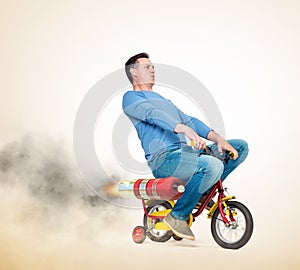 Cool man in casual wear rides a children`s bike with alternative rocket engine. On a light yellow background