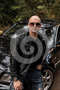 Cool man in sunglasses in style black jacket stands near black car on nature. Handsome guy travels by auto on beautiful sunny