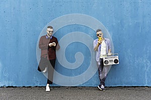 Cool men looking at mobile phone lean against blue wall - Couple of influencer guys listening music with vintage boombox stereo