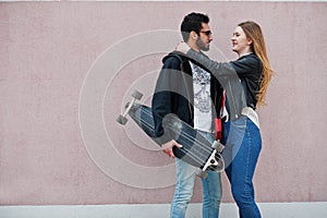 Cool multiracial couple posing against pink wall with longboard