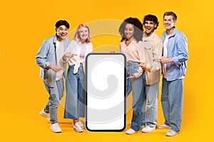 Cool offer. Happy diverse friends pointing at big blank smartphone screen over yellow background, mockup, full length