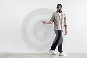 Cool offer. Happy young black guy pointing aside at free space, walking against white wall, full length portrait