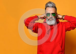 Cool old man with a gray beard is trying to put on a hood of a red sweatshirt hoodie on yellow in sunglasses