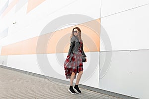 Cool positive young girl in red youth checkered dress in vintage leather jacket in trendy sunglasses in sneakers with bag travel