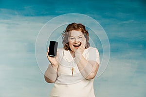 Cool red-haired plump woman in a white dress exults with a phone in her hands on a blue background photo