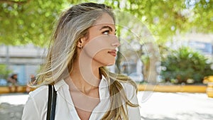 Cool and relaxed young blonde woman standing outdoors in a sunny park, giving a serious sideways glance - a captivating lifestyle