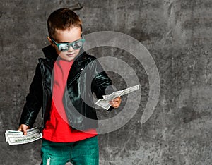 Cool rich kid boy in leather jacket, red t-shirt and stylish torn jeans is looking at bundles of dollars in his hands